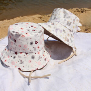 Two Baby Sunhats sit on a white towel. One floral hat sitting upright and the second hat leaning on the first at an angle. It has a dragon and knight on it. These are patterned hats. Small images cover the hats with a white background.